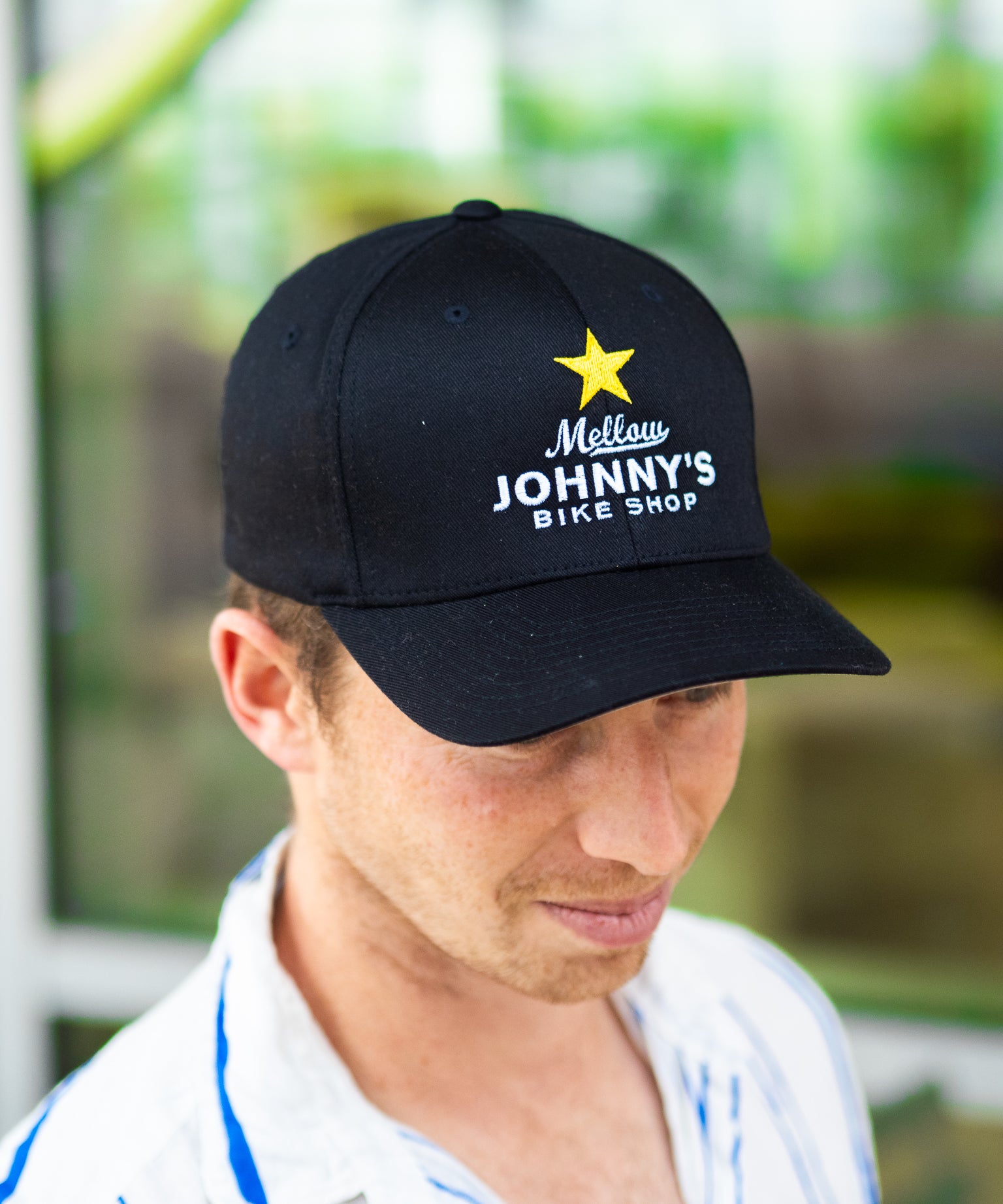 Man wearing a black cap with 'Mellow Johnny's Bike Shop' logo, blurred green background