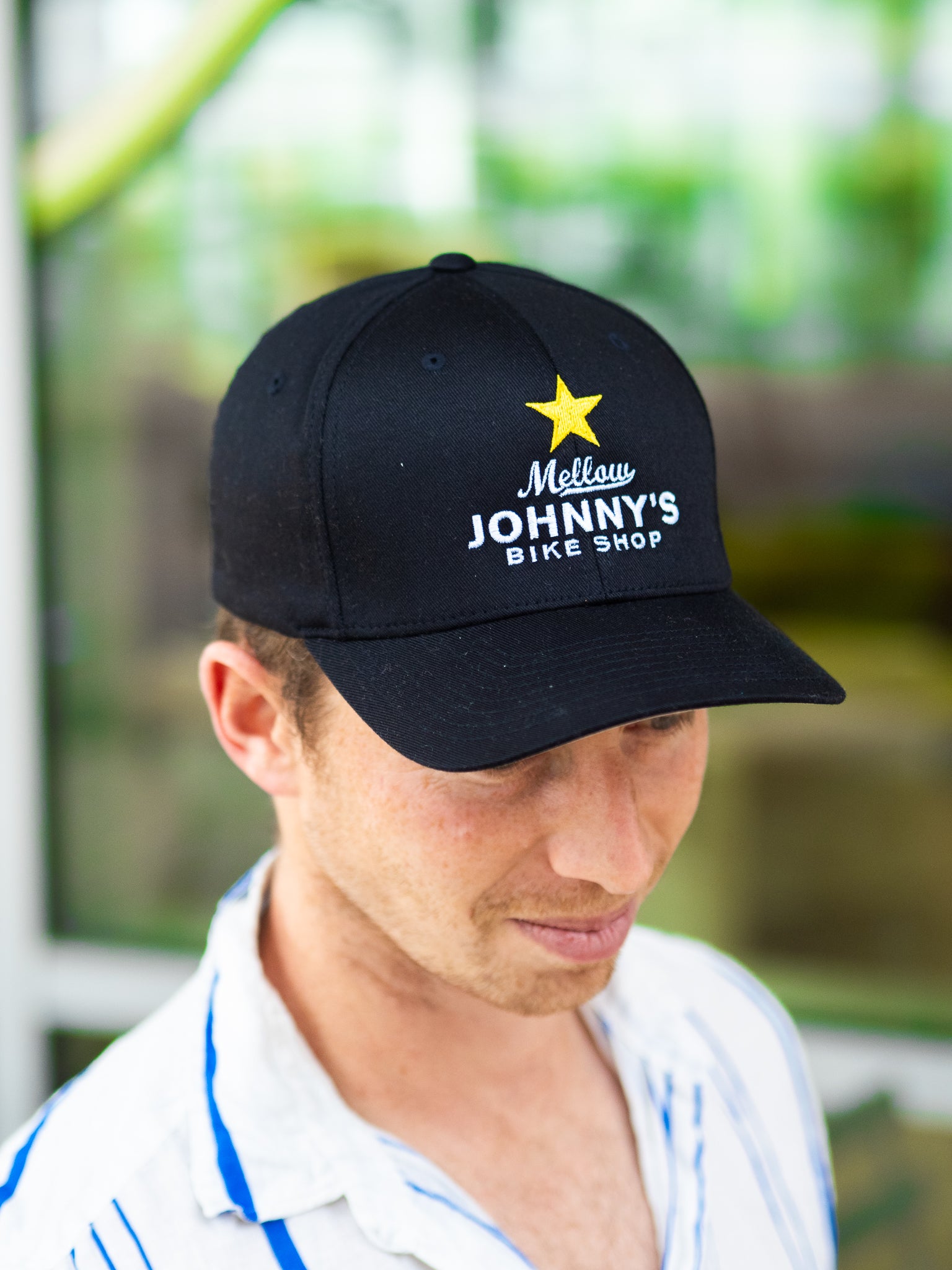 Man wearing a black cap with 'Mellow Johnny's Bike Shop' logo, blurred green background