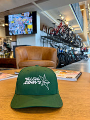 Green cap with 'Mellow Johnny's' logo on a table in a casual setting with bicycles and a TV in the background.
